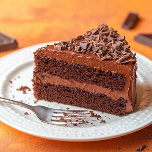 A slice of moist chocolate cake on a white, ridged plate with a silver fork